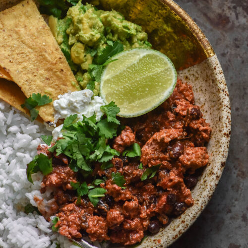 An aerial image of a speckled ceramic bowl filled with rice, low FODMAP vegan nomato chilli, guacamole, corn chips and a wedge of lime. The bowl sits on a speckled steel backdrop.