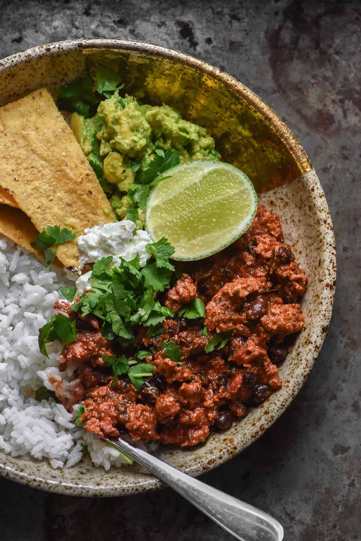 An aerial image of a speckled ceramic bowl filled with rice, low FODMAP vegan nomato chilli, guacamole, corn chips and a wedge of lime. The bowl sits on a speckled steel backdrop.