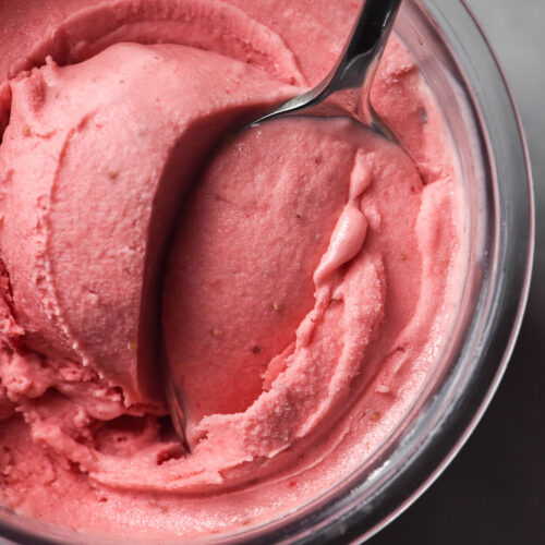 An aerial image of a tub of Ninja Creami sugar free strawberry ice cream on a white marble backdrop with a spoon sticking out of the tub