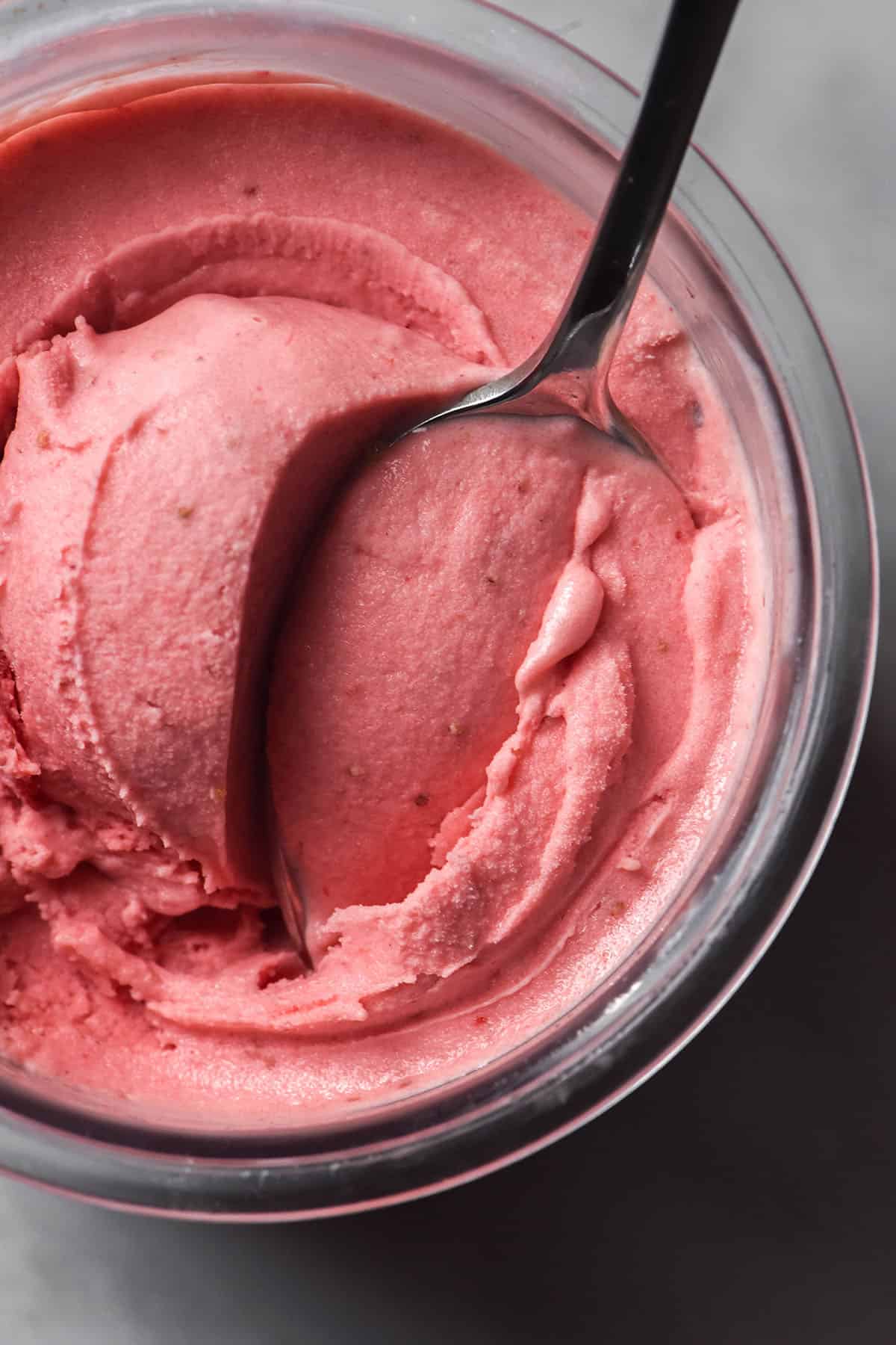 An aerial image of a tub of Ninja Creami sugar free strawberry ice cream on a white marble backdrop with a spoon sticking out of the tub