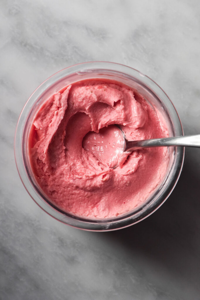 An aerial image of a Ninja Creami allulose strawberry ice cream tub on a white marble table. A spoon sits in the tub and sticks out to the right hand side.