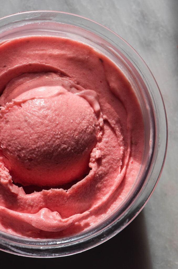 An aerial image of a Ninja Creami allulose strawberry ice cream tub on a white marble table. A scoop sits in the centre of the tub.