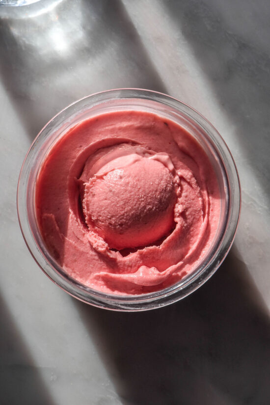 An aerial image of a tub of Ninja Creami sugar free strawberry ice cream on a sunlit white marble table. The tub has a scoop of ice cream in the centre and a sunlit water glass sits in the top left of the image.