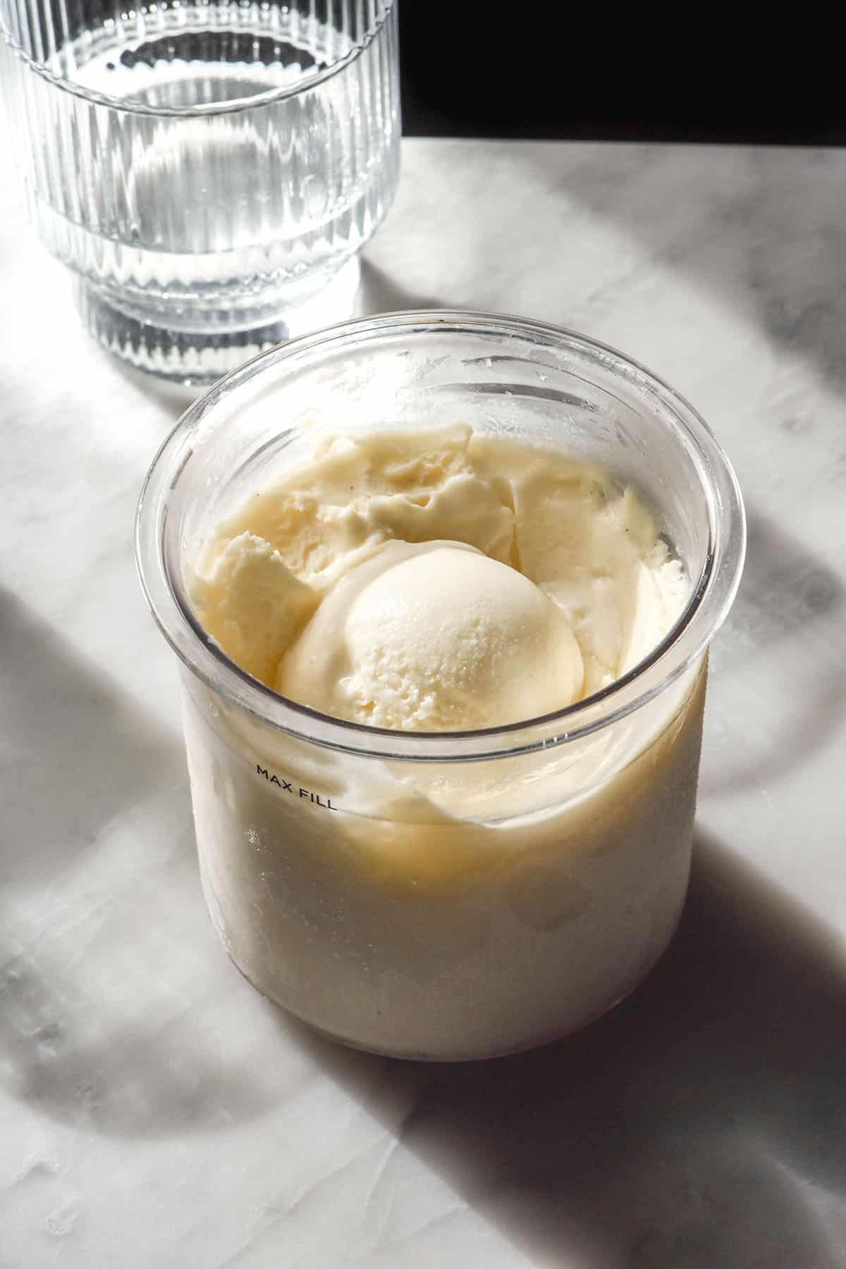 A side on image of a tub of Ninja Creami sugar free vanilla ice cream on a marble table in contrasting sunlight. A scoop of ice cream sits in the centre of the tub and a sunlit glass of water sits off to the left of the image.