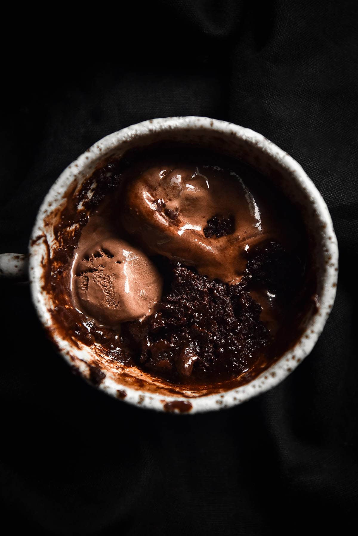 A dark and moody aerial close up image of a gluten free Nutella mug cake in a white speckled ceramic cup atop a black backdrop