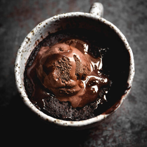 A moody image of a gluten free chocolate nutella mug cake in a white speckled ceramic mug. The cake is topped with a melting scoop of chocolate ice cream and the mug sits atop a mottled steel backdrop.