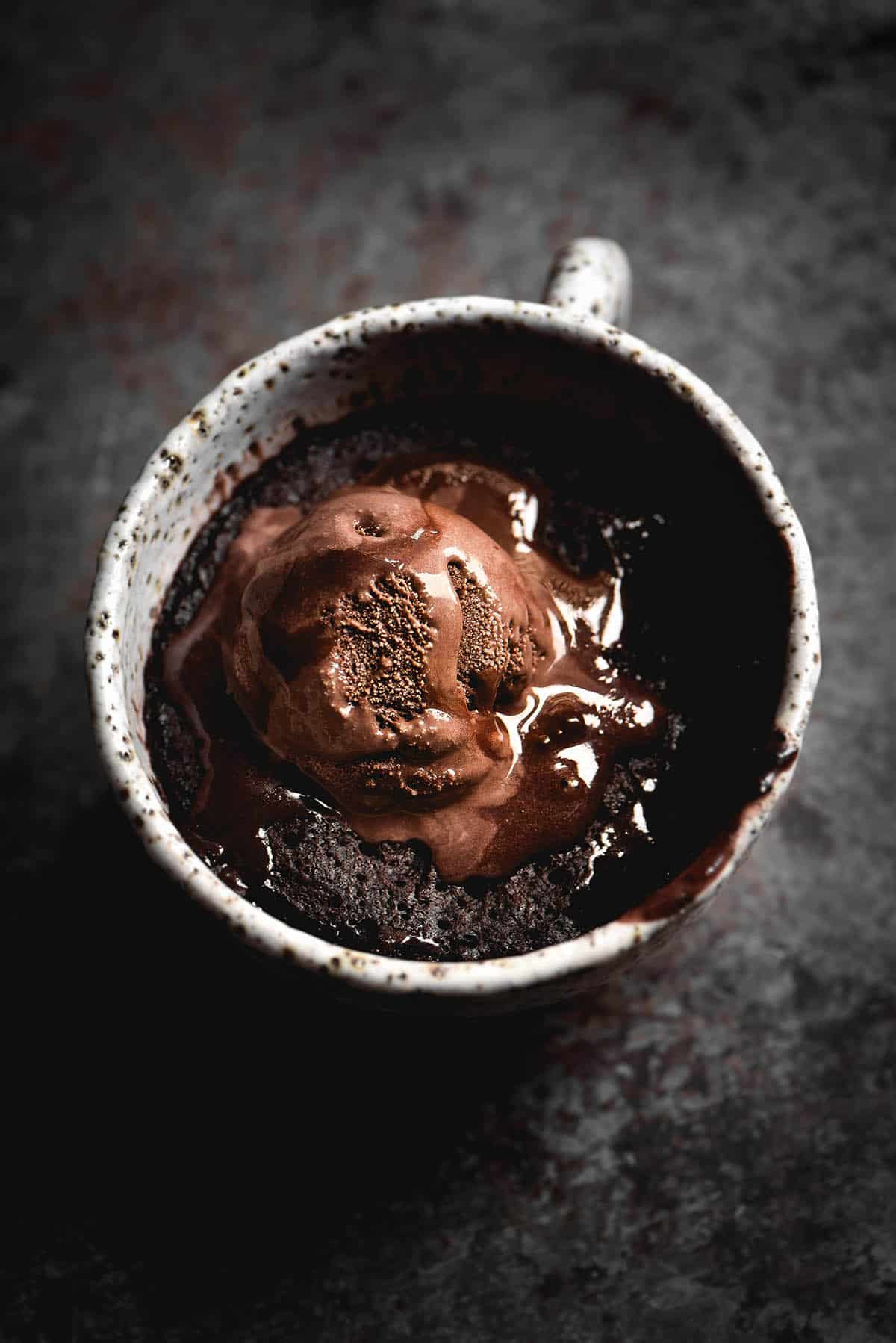 A moody image of a gluten free chocolate nutella mug cake in a white speckled ceramic mug. The cake is topped with a melting scoop of chocolate ice cream and the mug sits atop a mottled steel backdrop.