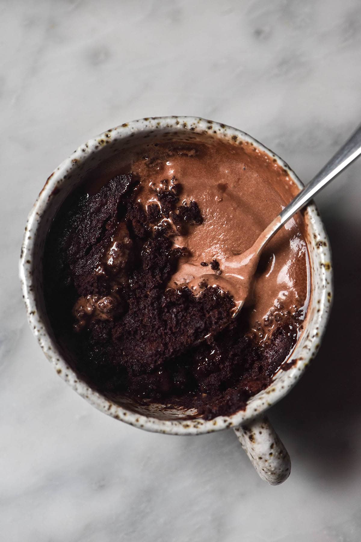 An aerial image of a gluten free Nutella mug cake topped with chocolate ice cream in a white ceramic mug atop a white marble table. A spoon sticks into the mug cake from the right side of the image.
