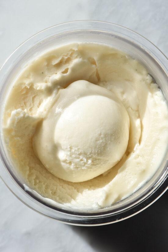 An aerial macro image of a tub of sugar free vanilla Ninja Creami ice cream on a white marble table. The ice cream has a scoop on the centre of the tub.