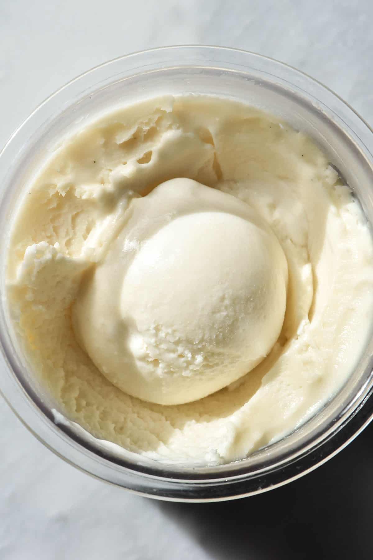 An aerial macro image of a tub of sugar free vanilla Ninja Creami ice cream on a white marble table. The ice cream has a scoop on the centre of the tub.