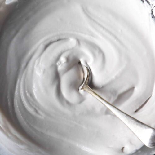 An aerial image of a bowl of homemade coconut yoghurt atop a white marble table. A spoon sits in the centre of the bowl.