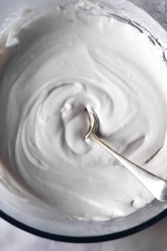 An aerial image of a bowl of homemade coconut yoghurt atop a white marble table. A spoon sits in the centre of the bowl.