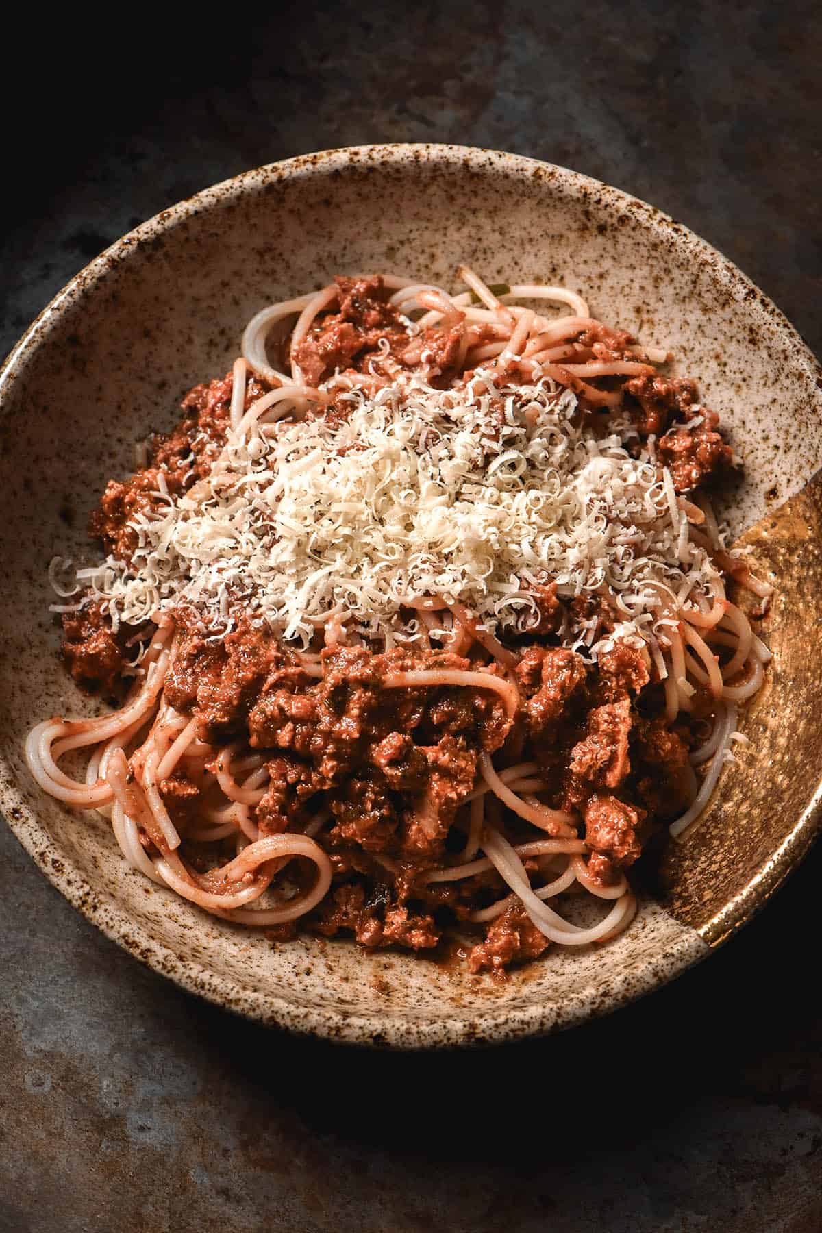 An aerial image of a speckled ceramic bowl filled with low FODMAP spaghetti bolognese and topped with plenty of grated parmesan. The bowl sits atop a dark steel backdrop.