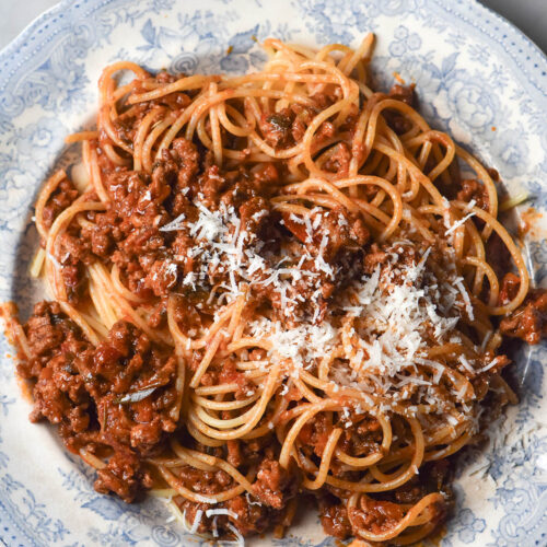 An aerial image of a blue and white ceramic plate topped with low FODMAP spaghetti bolognese. The bolognese is topped with plenty of parmesan cheese and the plate sits atop a white marble table.