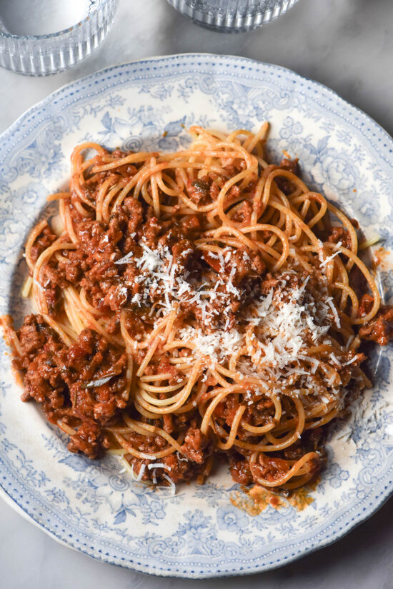 An aerial image of a blue and white ceramic plate topped with low FODMAP spaghetti bolognese. The bolognese is topped with plenty of parmesan cheese and the plate sits atop a white marble table.