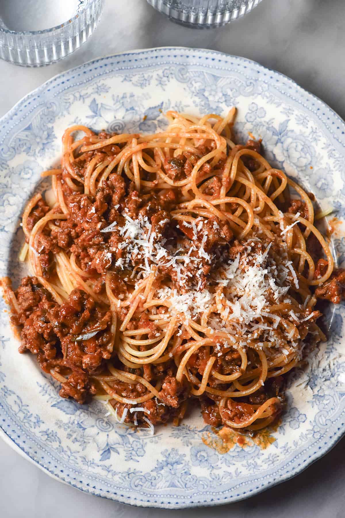 An aerial image of a blue and white ceramic plate topped with low FODMAP spaghetti bolognese. The bolognese is topped with plenty of parmesan cheese and the plate sits atop a white marble table.