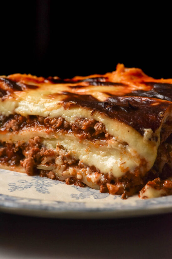 A moody macro image of a melty slice of low FODMAP lasagne on a blue and white patterned plate against a black backdrop