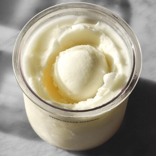An aerial image of a tub of Ninja Creami lemon sorbet on a sunlit white marble table. The tub has a scoop of sorbet in the middle.