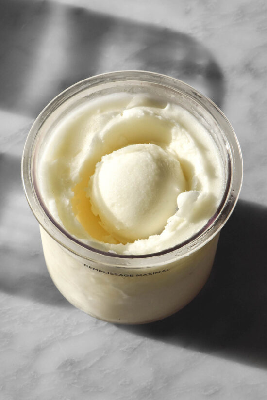 An aerial image of a tub of Ninja Creami lemon sorbet on a sunlit white marble table. The tub has a scoop of sorbet in the middle.
