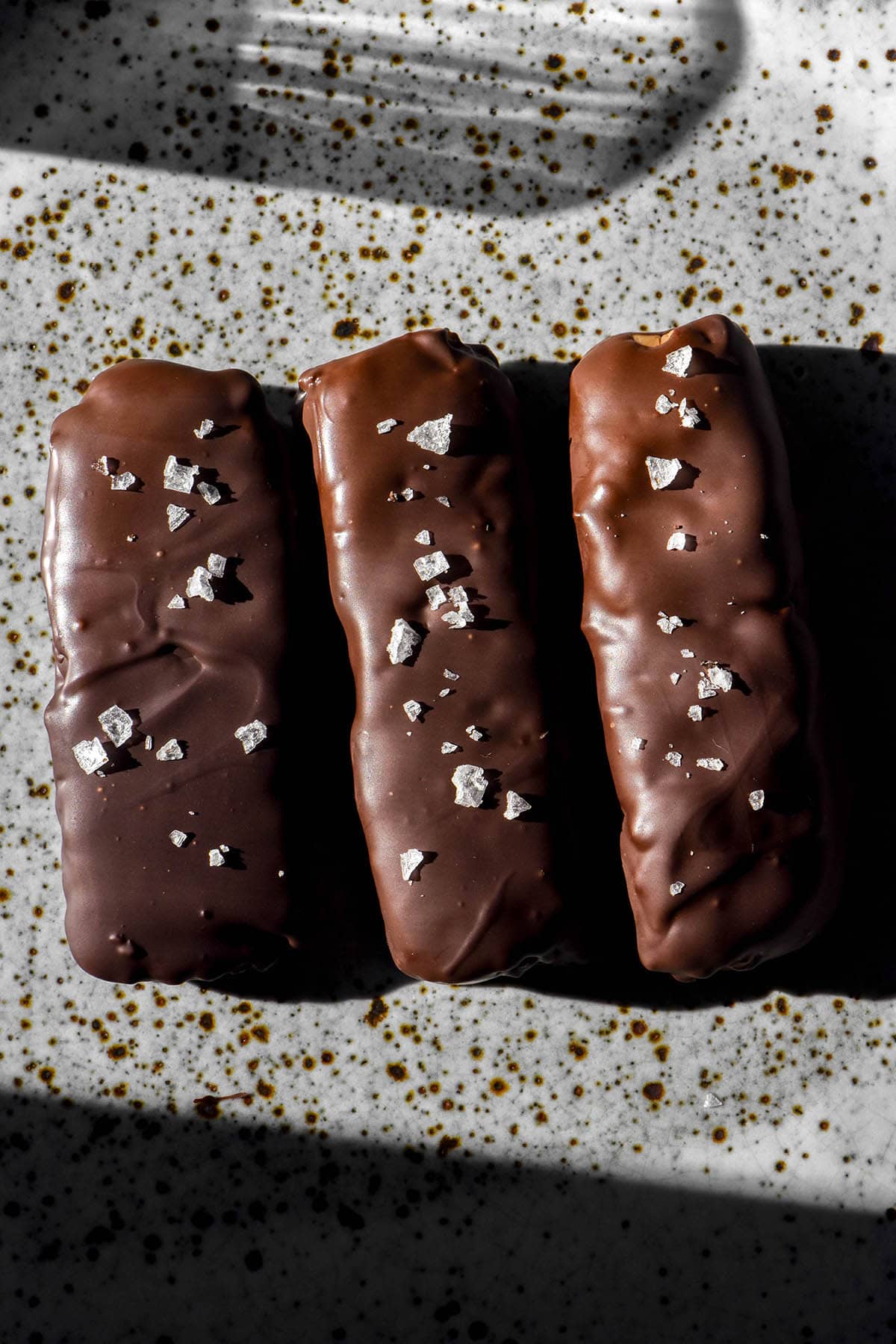 An aero macro image of three chocolate coated protein crisp bars on a white speckled ceramic plate topped with sea salt flakes. The plate sits in contrasting sunlight and the reflection of a water glass sits in the top of the image.