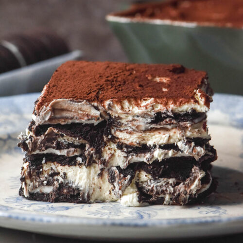 A side on slice of gluten free Oreo Tiramisu on a white ceramic plate against a brown backdrop. The container of Tiramisu and extra Oreo biscuits sit in the background.