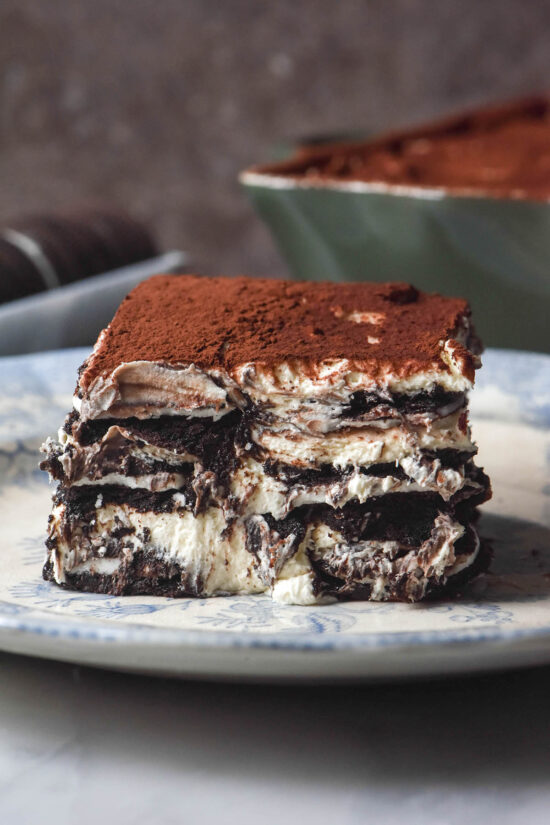 A side on slice of gluten free Oreo Tiramisu on a white ceramic plate against a brown backdrop. The container of Tiramisu and extra Oreo biscuits sit in the background.
