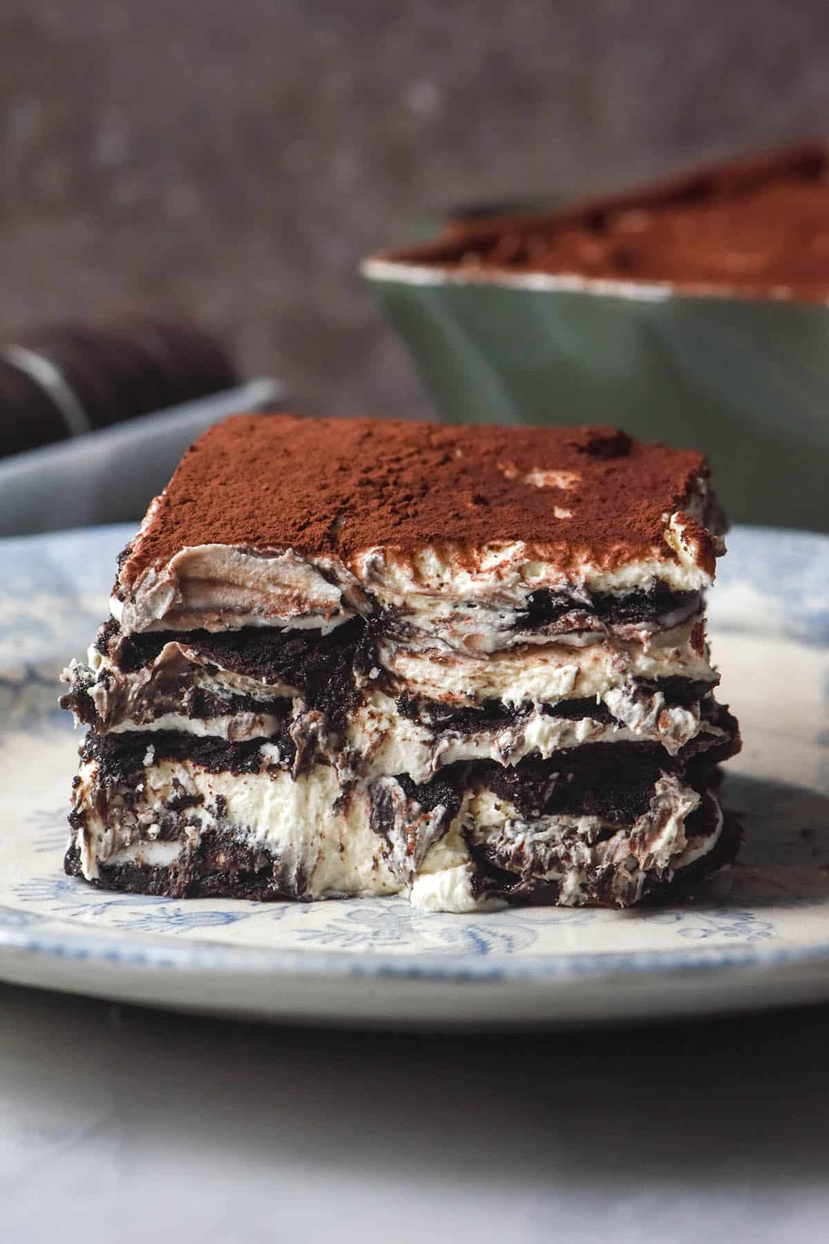A side on slice of gluten free Oreo Tiramisu on a white ceramic plate against a brown backdrop. The container of Tiramisu and extra Oreo biscuits sit in the background.