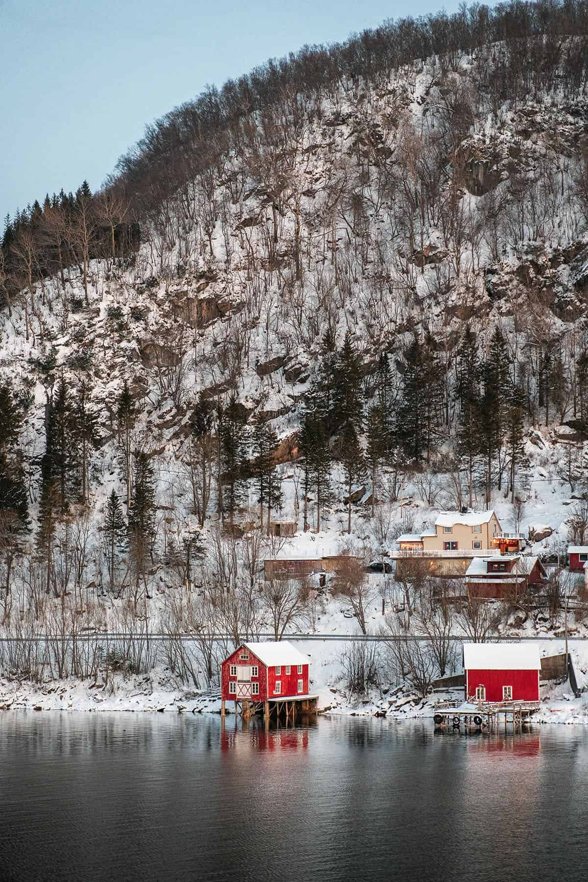 An image of rorbu on the water in coastal Norway. Snow has started to blanket the trees on the hillside and the sky is blue