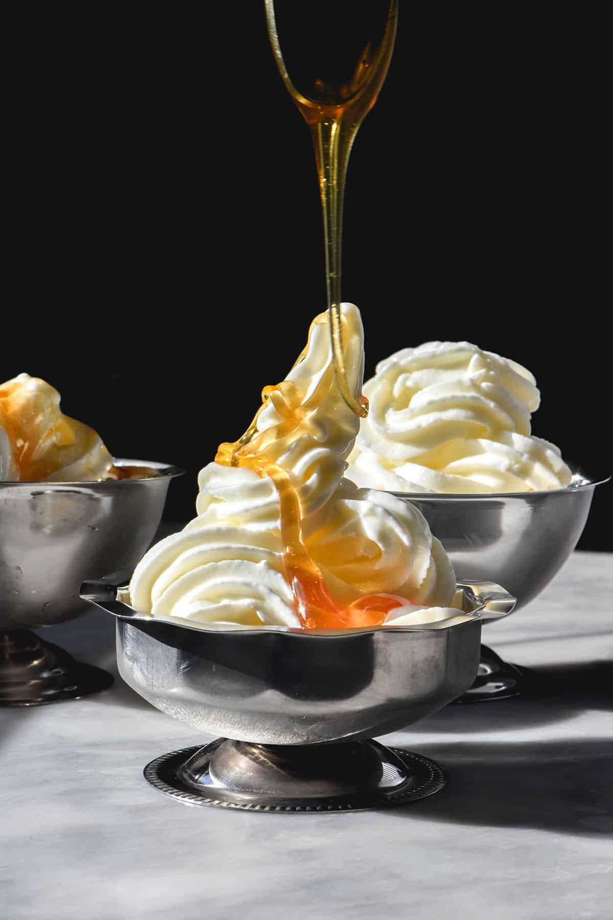 A side on image of 3 silver dessert coupes filled with frozen yogurt soft serve. The coupes sit on a white table against a dark backdrop. Honey is being drizzled down onto the central coupe of frozen yoghurt
