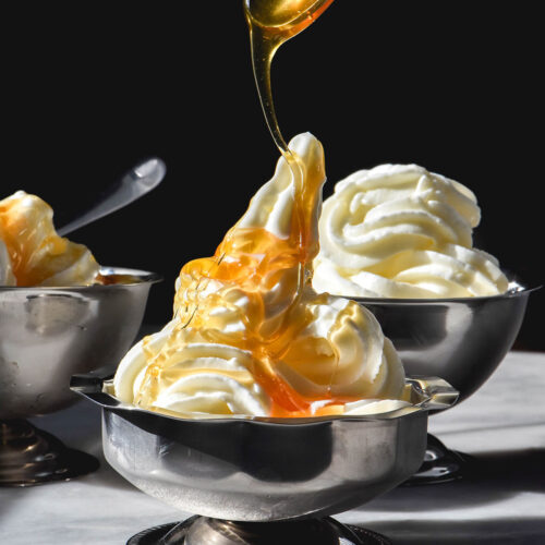A side on image of 3 silver dessert coupes filled with frozen yogurt soft serve. The coupes sit on a white table against a dark backdrop. Honey is being drizzled down onto the central coupe of frozen yoghurt