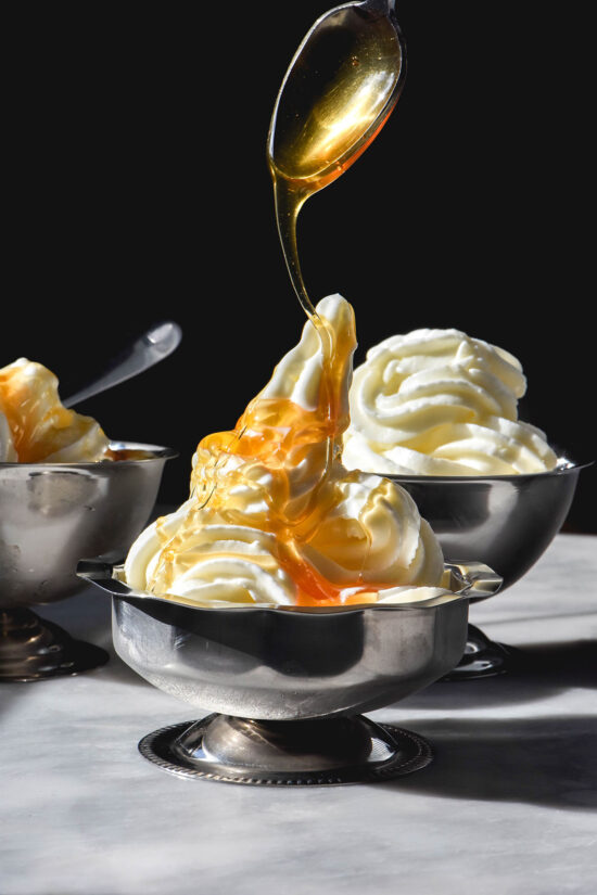 A side on image of 3 silver dessert coupes filled with frozen yogurt soft serve. The coupes sit on a white table against a dark backdrop. Honey is being drizzled down onto the central coupe of frozen yoghurt
