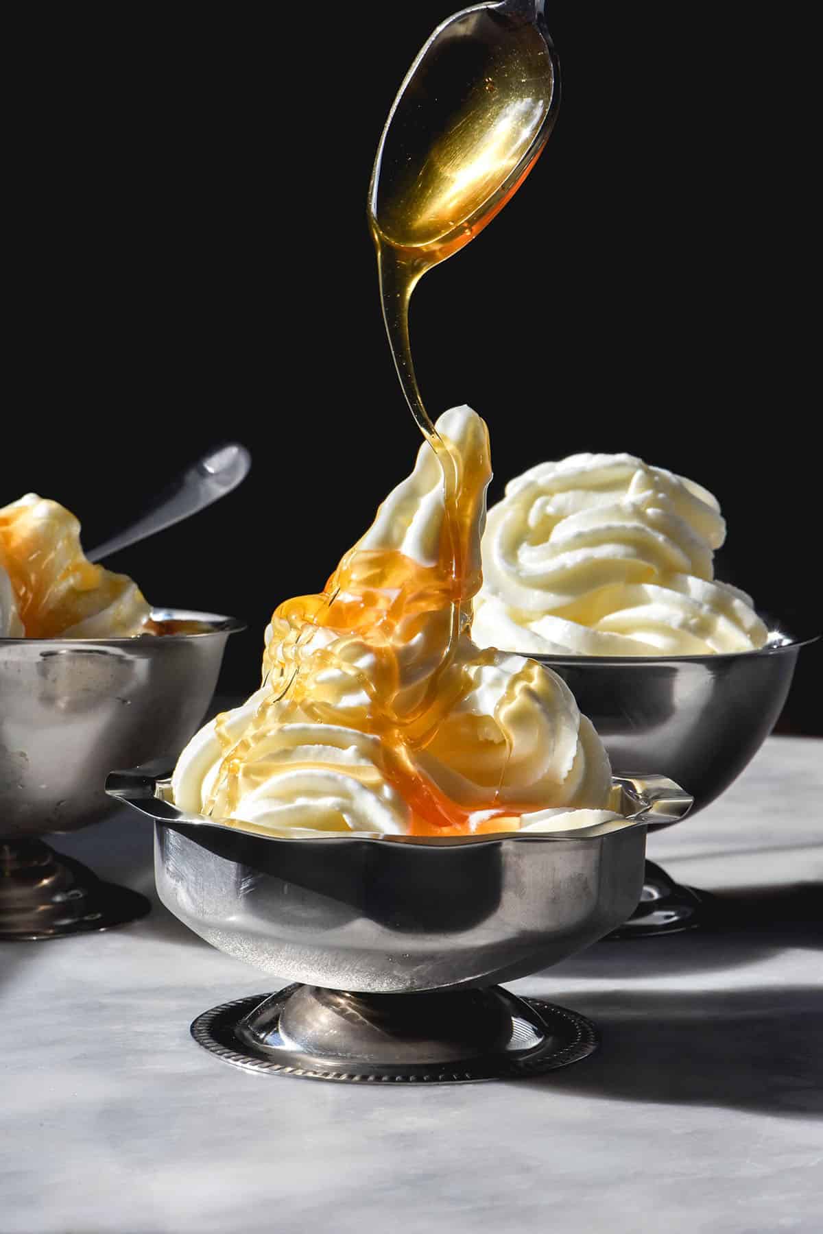 A side on image of 3 silver dessert coupes filled with frozen yogurt soft serve. The coupes sit on a white table against a dark backdrop. Honey is being drizzled down onto the central coupe of frozen yoghurt