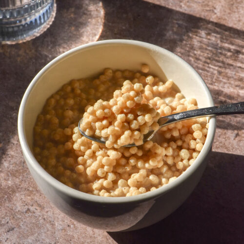 An aerial sunlit image of a small white ceramic bowl filled with protein crisp cereal. A spoon dips into the bowl from the right side of the image and a sunlit water glass sits on the left side of the image.