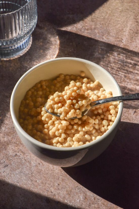 An aerial sunlit image of a small white ceramic bowl filled with protein crisp cereal. A spoon dips into the bowl from the right side of the image and a sunlit water glass sits on the left side of the image.