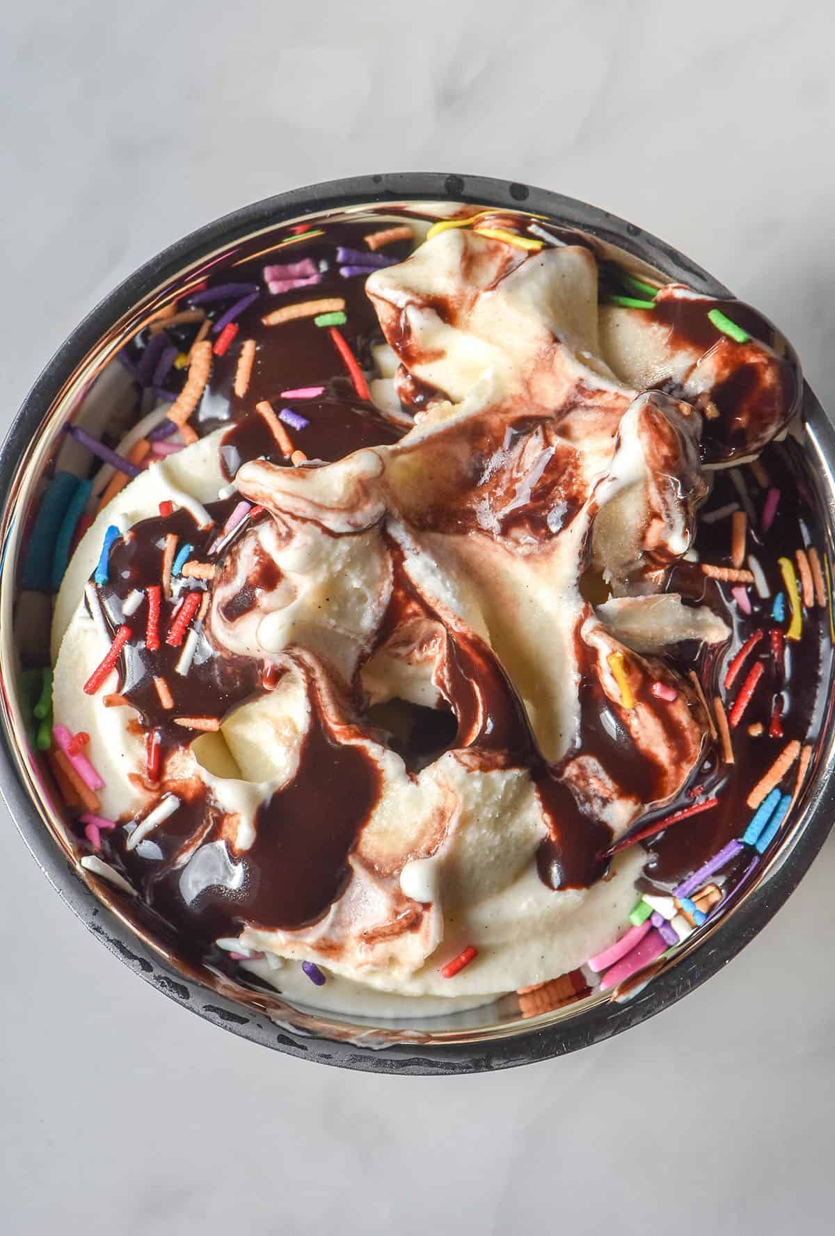 An aerial image of a melting vanilla soft serve topped with brown butter hot fudge sauce and rainbow sprinkles. The dessert sits in a silver coupe on a white marble table
