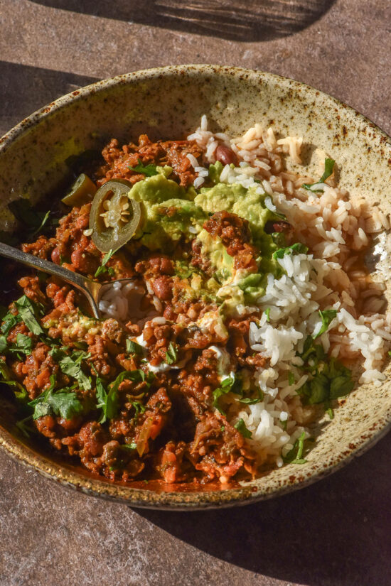 A sunlit image of a bowl of low FODMAP chilli con carne on a brown backdrop. The chilli is served with rice and casually topped with avocado, coriander and pickled jalapeno