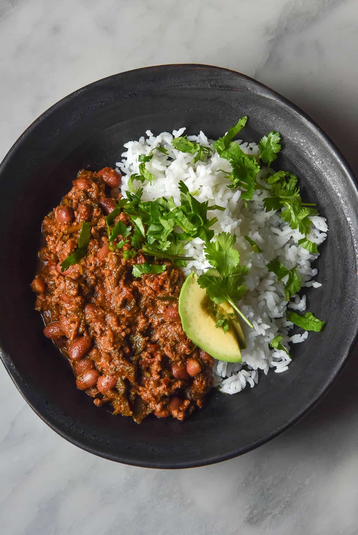 An aerial image of a dark blue bowl filled with low FODMAP chilli con carne on a white marble table. The chilli is served with white rice, avocado and fresh coriander