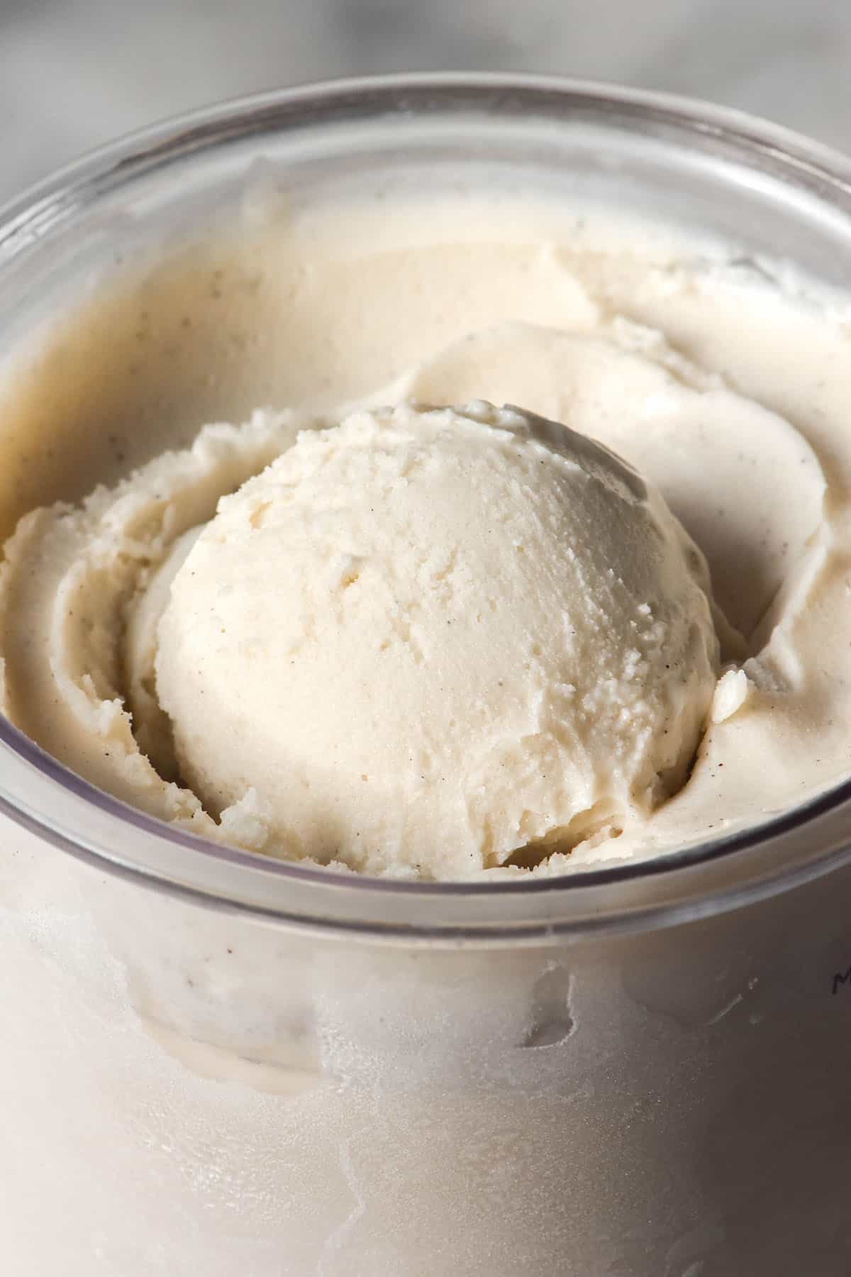 A close up macro image of a tub of Ninja Creami cashew ice cream on a white marble table. There is a scoop of ice cream in the centre of the tub.