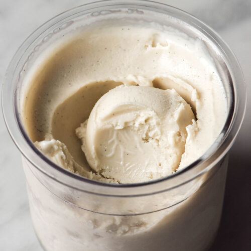 An aerial image of a tub of Ninja Creami cashew ice cream on a white marble table. A scoop sits in the centre of the tub and a spoonful has been removed, revealing the creamy texture