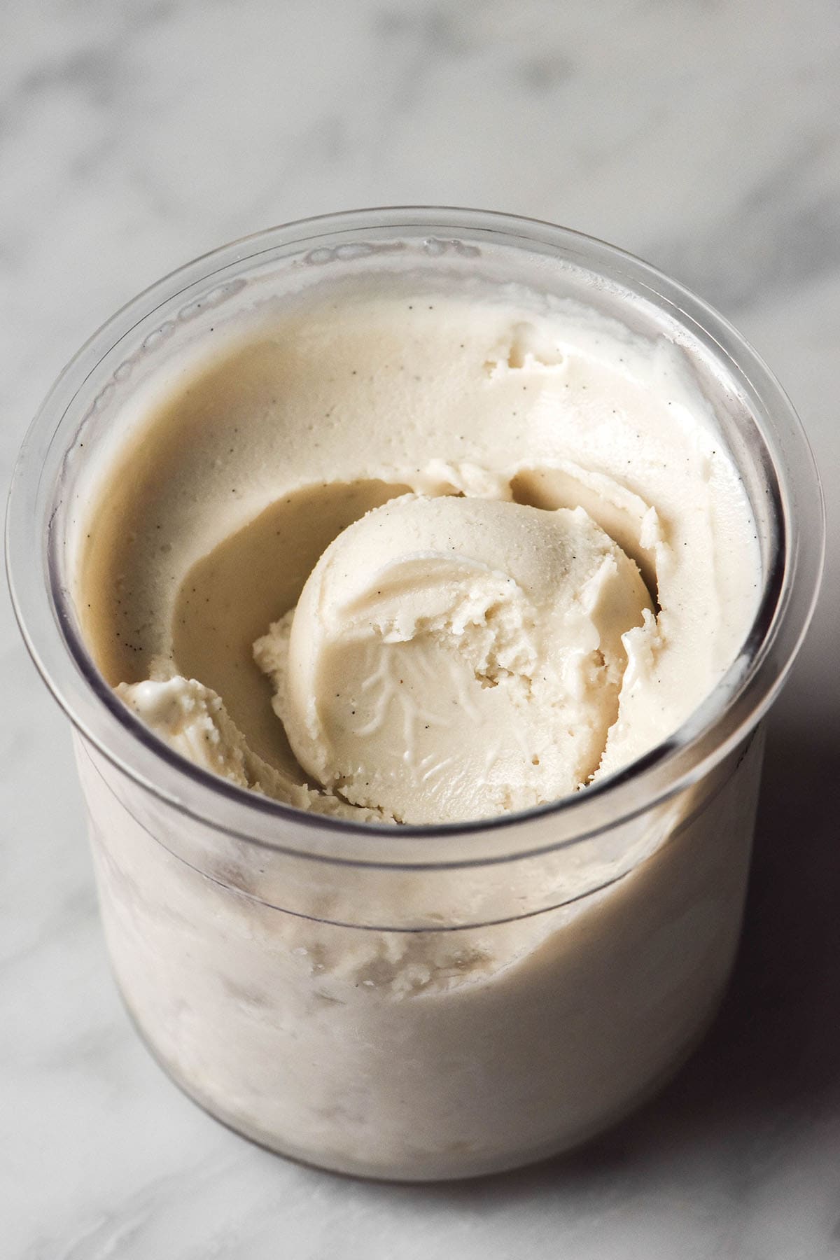 An aerial image of a tub of Ninja Creami cashew ice cream on a white marble table. A scoop sits in the centre of the tub and a spoonful has been removed, revealing the creamy texture