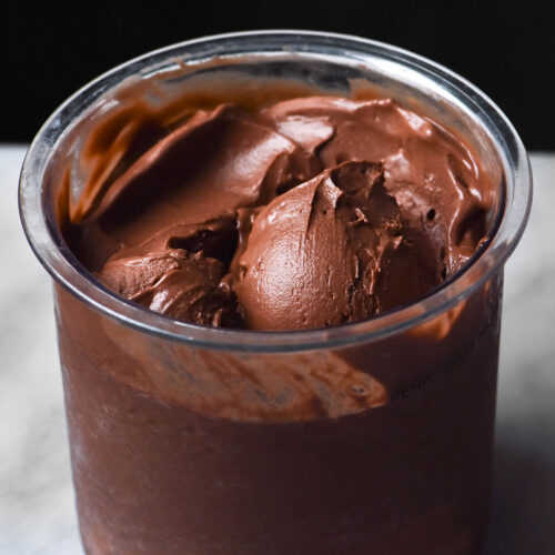 A side on image of a tub of Ninja Creami vegan chocolate ice cream on a white marble table against a dark backdrop. A scoop of ice cream sits in the centre of the tub.