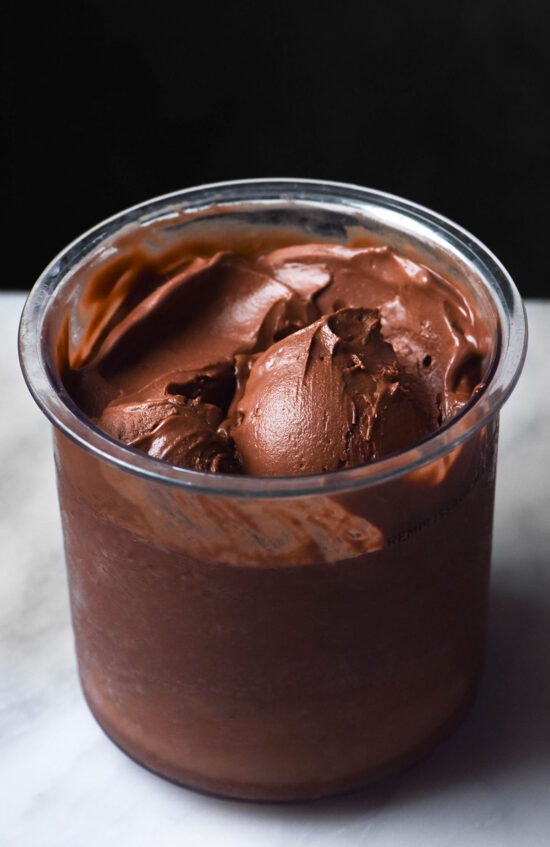 A side on image of a tub of Ninja Creami vegan chocolate ice cream on a white marble table against a dark backdrop. A scoop of ice cream sits in the centre of the tub.