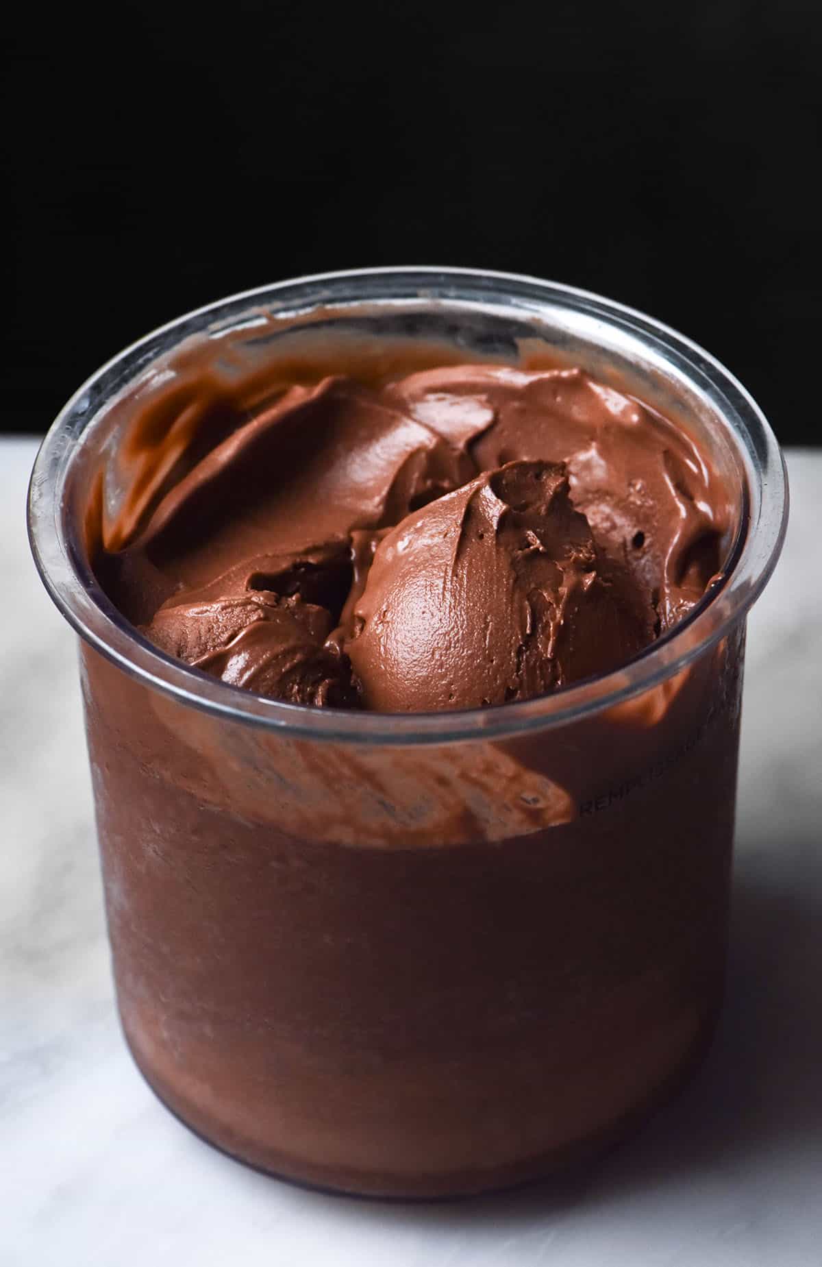 A side on image of a tub of Ninja Creami vegan chocolate ice cream on a white marble table against a dark backdrop. A scoop of ice cream sits in the centre of the tub.