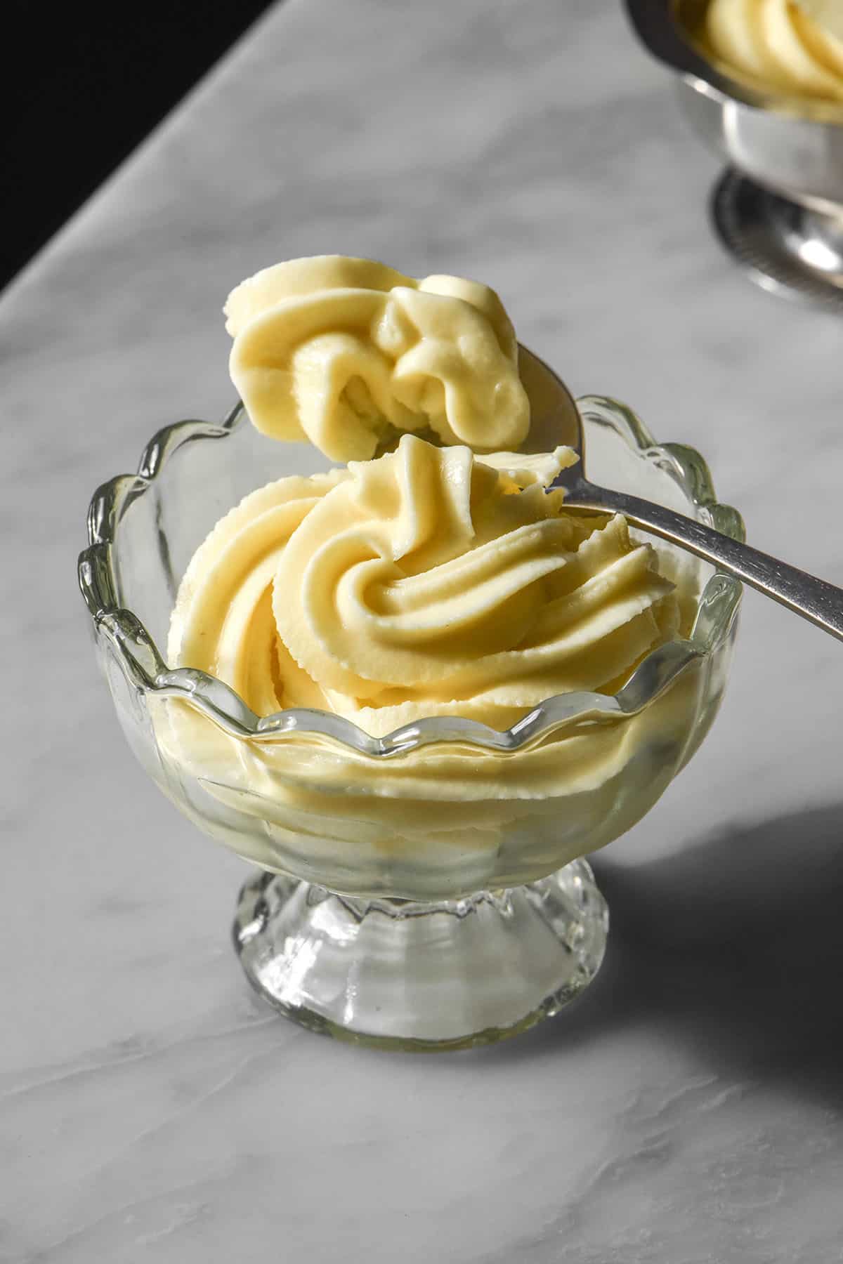 An image of Ninja Swirl Dole whip in a glass scalloped dessert cup on a white marble table. A spoon dips into the ice cream from the right side of the image.