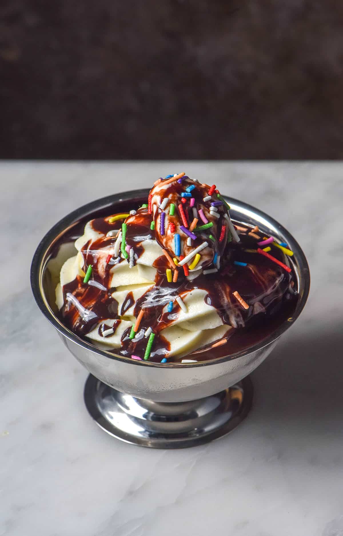 A side on image of a silver dessert coupe on a white marble table. The dessert coupe is filled with Ninja Swirl vanilla soft serve and topped with hot fudge sauce and rainbow sprinkles