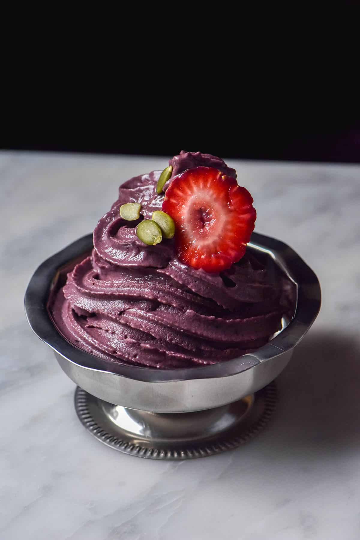 A silver dessert coupe filled with Ninja Swirl açai soft serve on a white marble table against a dark backdrop. The açai is topped with strawberries and pumpkin seeds.
