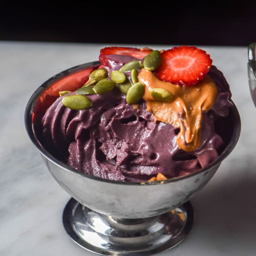 A silver dessert coupe filled with Ninja Swirl açai soft serve on a white marble table against a dark backdrop. The açai is topped with peanut butter, strawberries and pumpkin seeds.