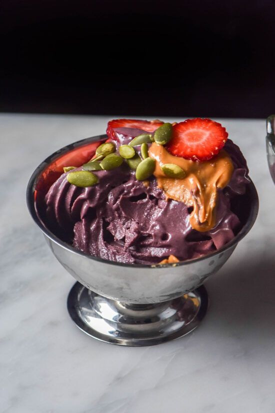 A silver dessert coupe filled with Ninja Swirl açai soft serve on a white marble table against a dark backdrop. The açai is topped with peanut butter, strawberries and pumpkin seeds.