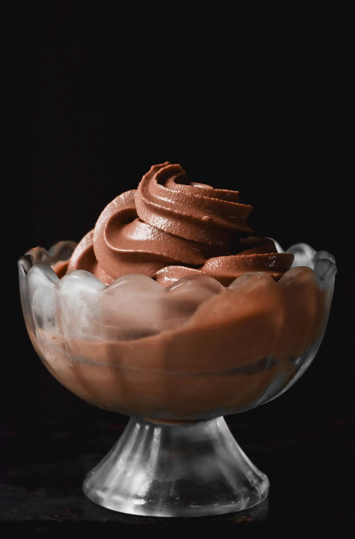 A moody image of a glass scalloped dessert coupe filled with Ninja Swirl chocolate soft serve against a dark backdrop