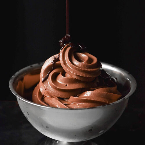 A side on moody image of a frosted silver dessert coupe filled with Ninja Swirl chocolate soft serve. A drizzle of brown butter hot fudge sauce swirls down from the top of the image